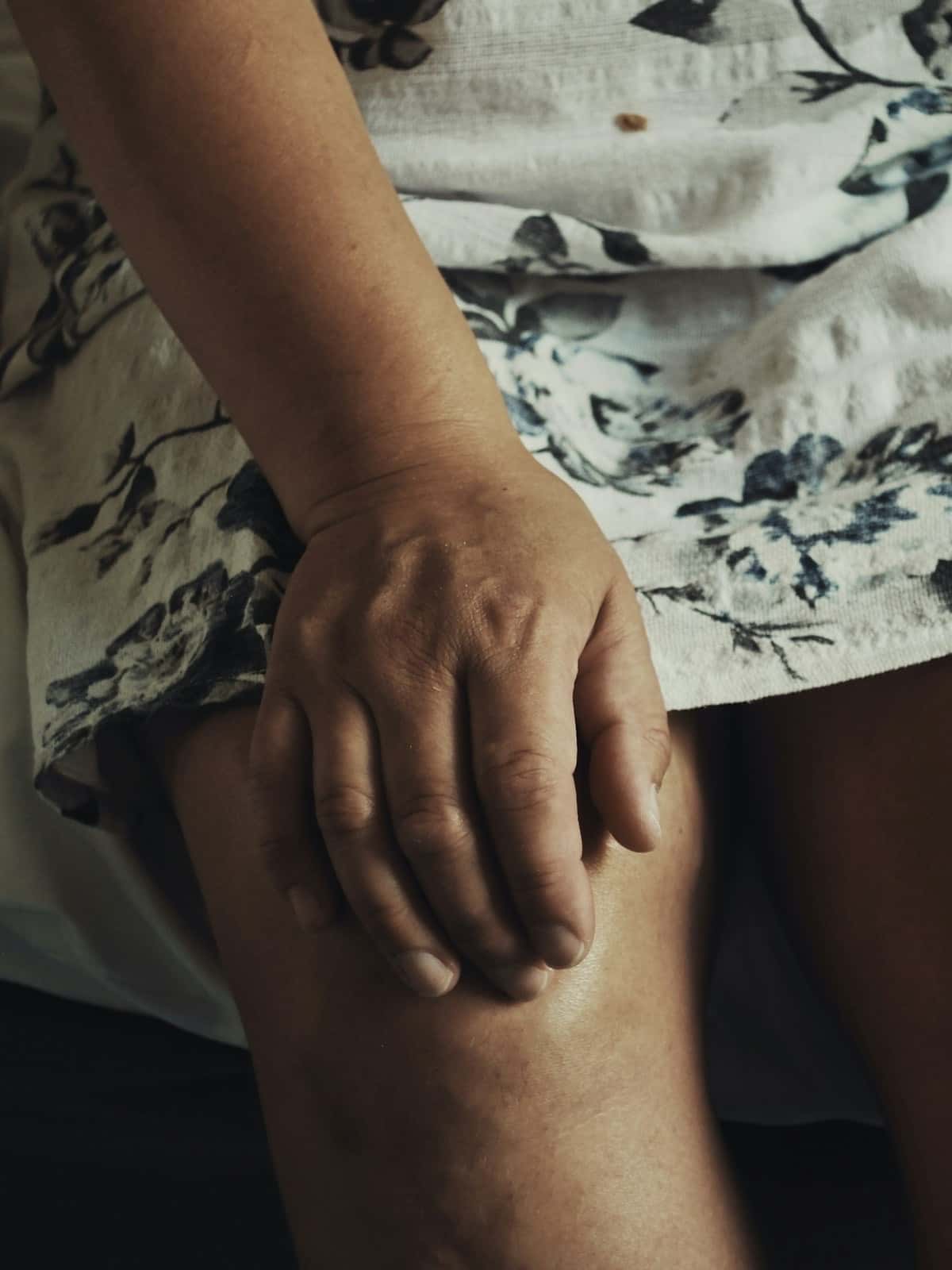 Close-up of a woman seated on a bed in a floral dress pressing her hand against her bare knee.