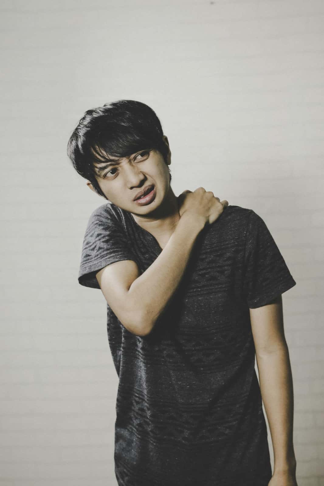 A young man in a grey t-shirt grimaces while gripping his right shoulder with his left hand against a plain white wall.