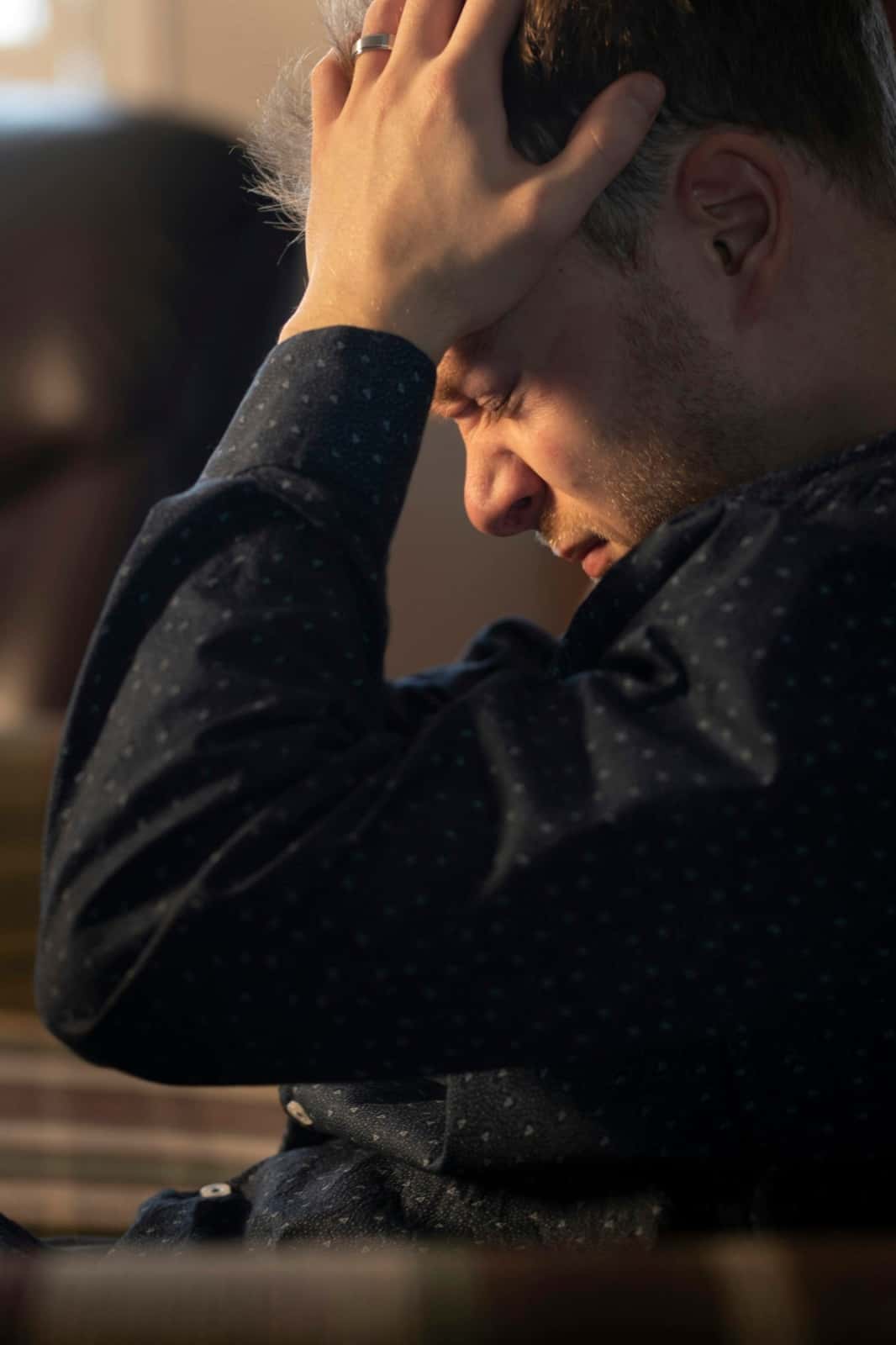 A man in a dark patterned shirt sits indoors with his eyes squeezed shut and one hand pressed to his forehead, lit by warm side light.