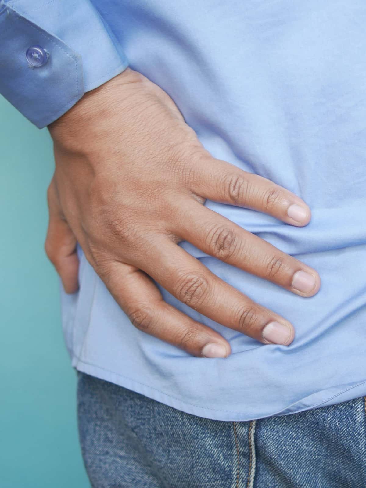 Close-up side view of a man in a light blue shirt and jeans pressing his hand against his hip in discomfort.