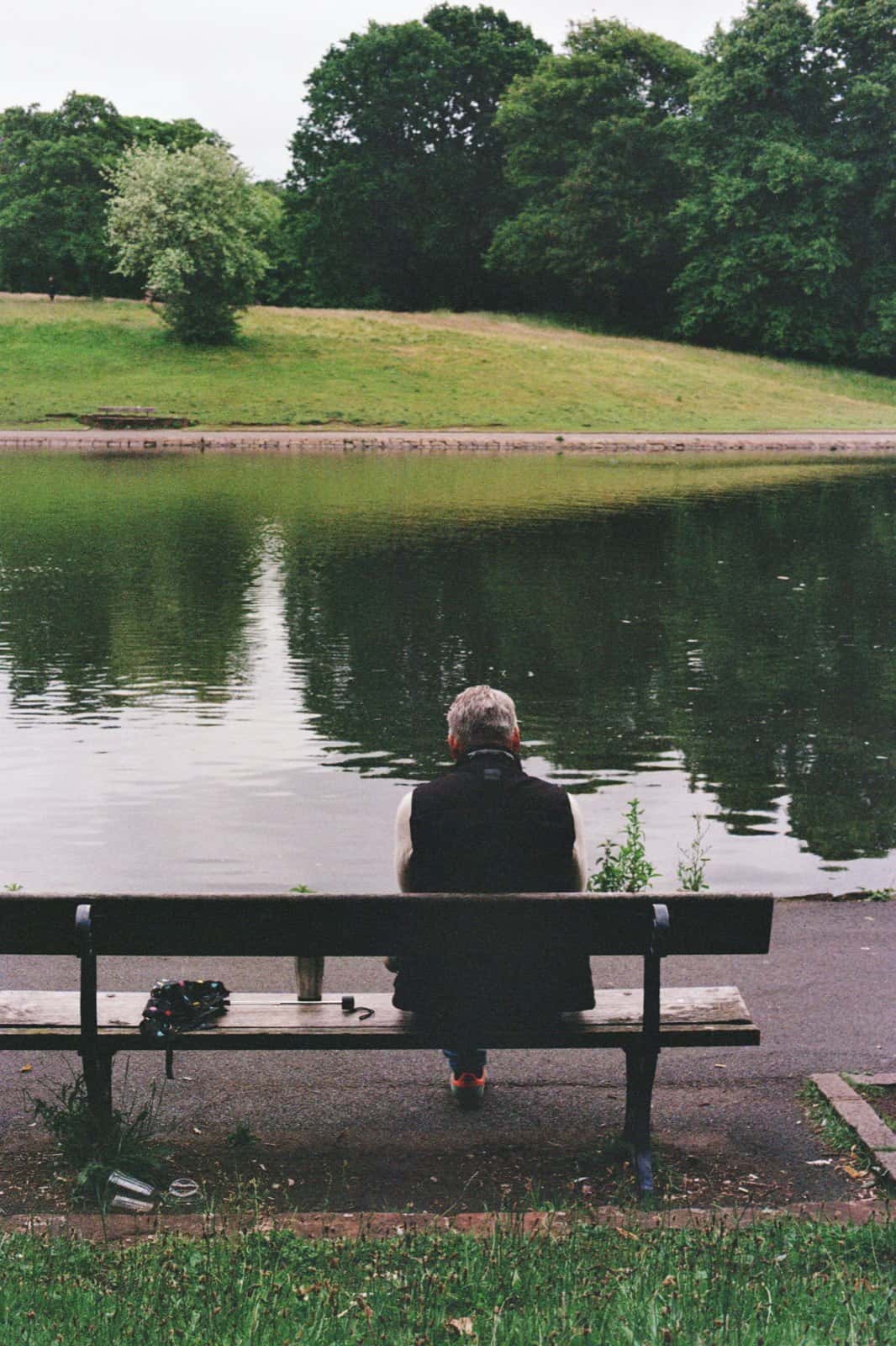A grey-haired man in a dark vest sits alone on a wooden park bench, seen from behind, resting and looking out over a calm pond and green hillside.