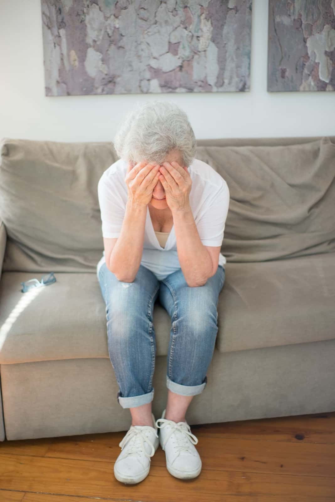 An older woman with gray hair sits on the edge of a beige sofa in jeans and a white shirt, head bowed with both hands covering her face.