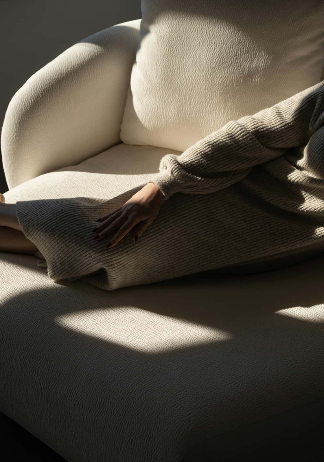 A person in an oversized ribbed knit sweater sits curled on a cream armchair with one hand resting on their lap in a patch of afternoon sunlight, face out of frame.