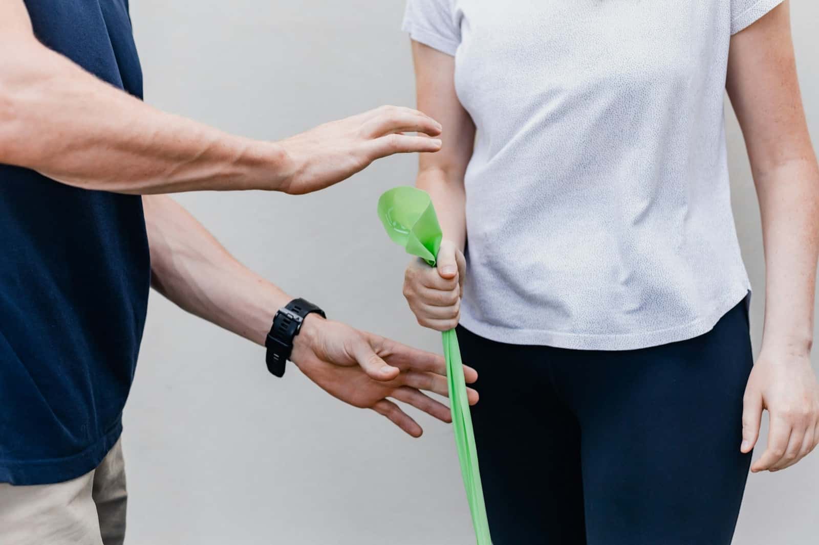 A physical therapist's hands guide a patient's arm as she pulls a green resistance band against a plain wall.
