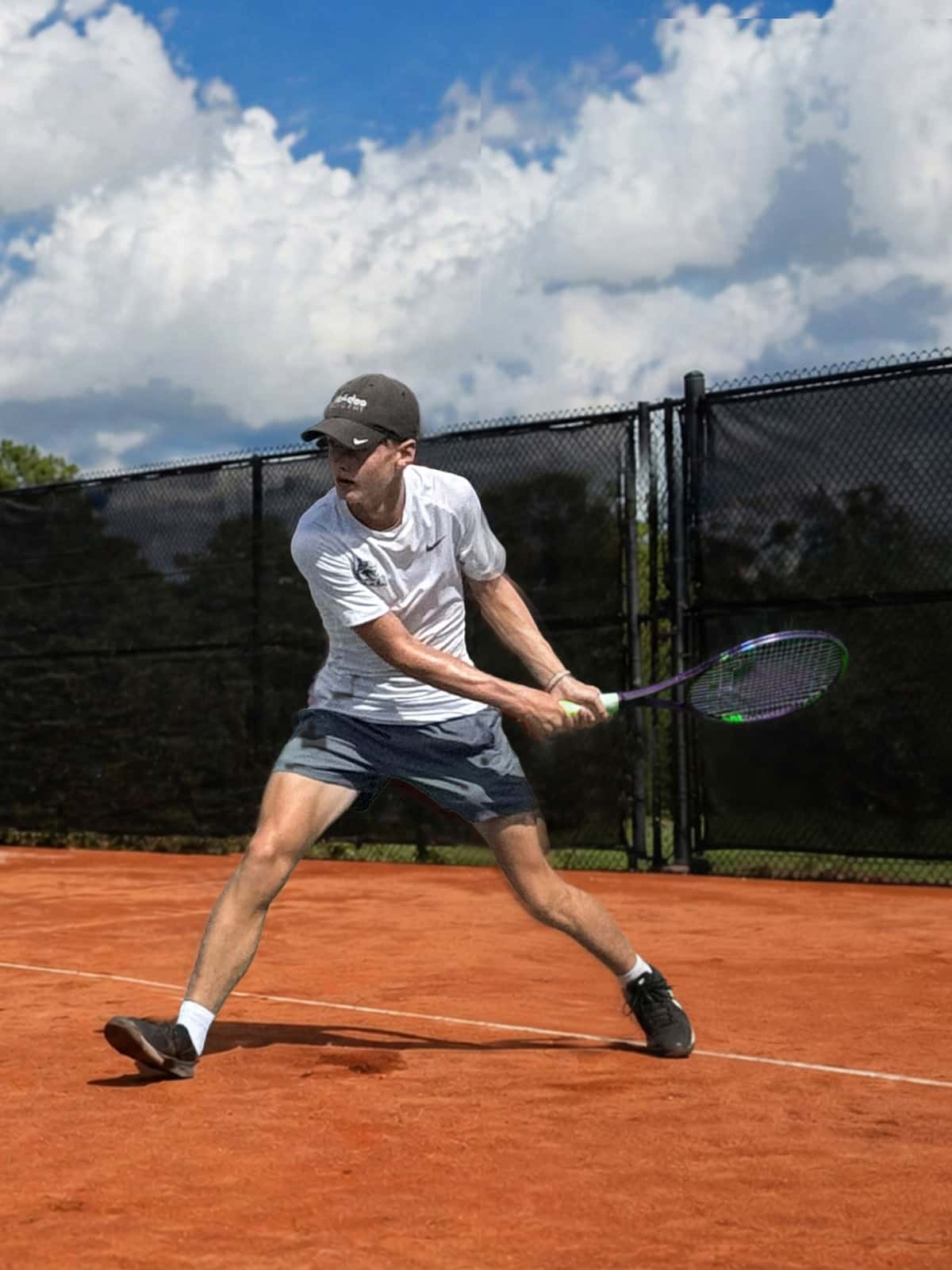 A young man in athletic wear and a dark cap swings a tennis racquet with both hands on an outdoor clay court, feet planted wide.