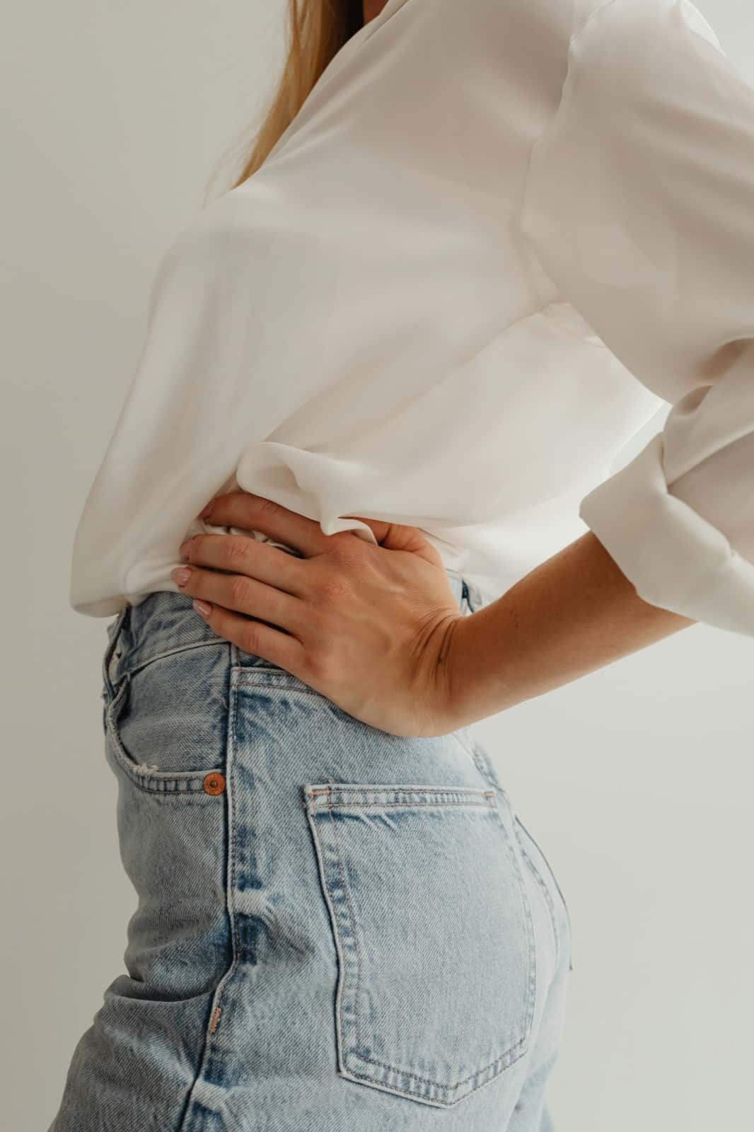 A woman in a white blouse and light-wash jeans stands in profile with one hand pressed against her lower back.