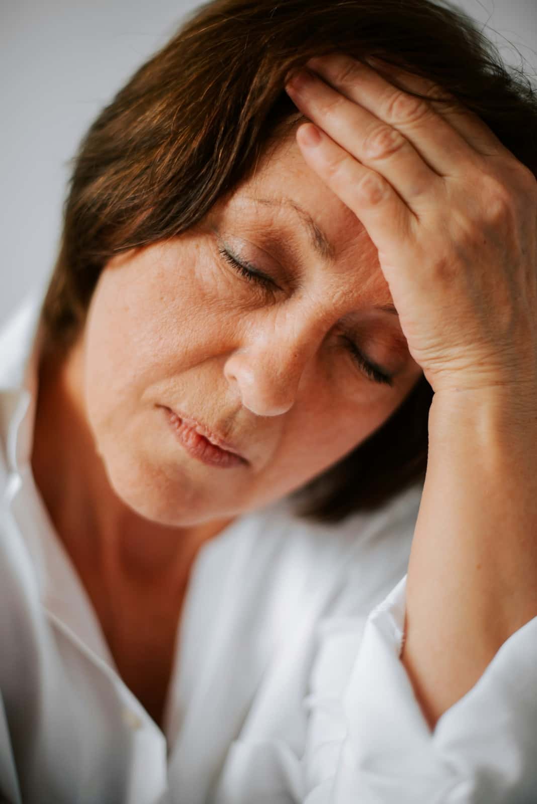 A woman in a white shirt with her eyes closed presses her hand against her temple and forehead in soft natural light.