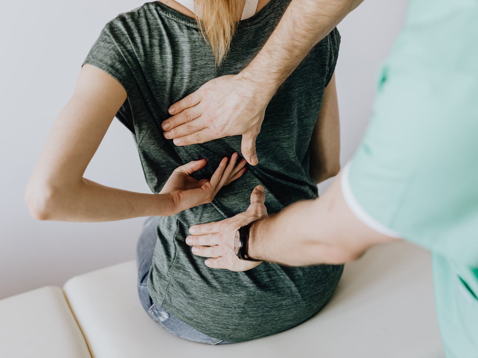 A clinician in mint-green scrubs presses one hand against the mid-back and the other against the lower back of a woman seated upright and fully clothed on the edge of a padded exam table.