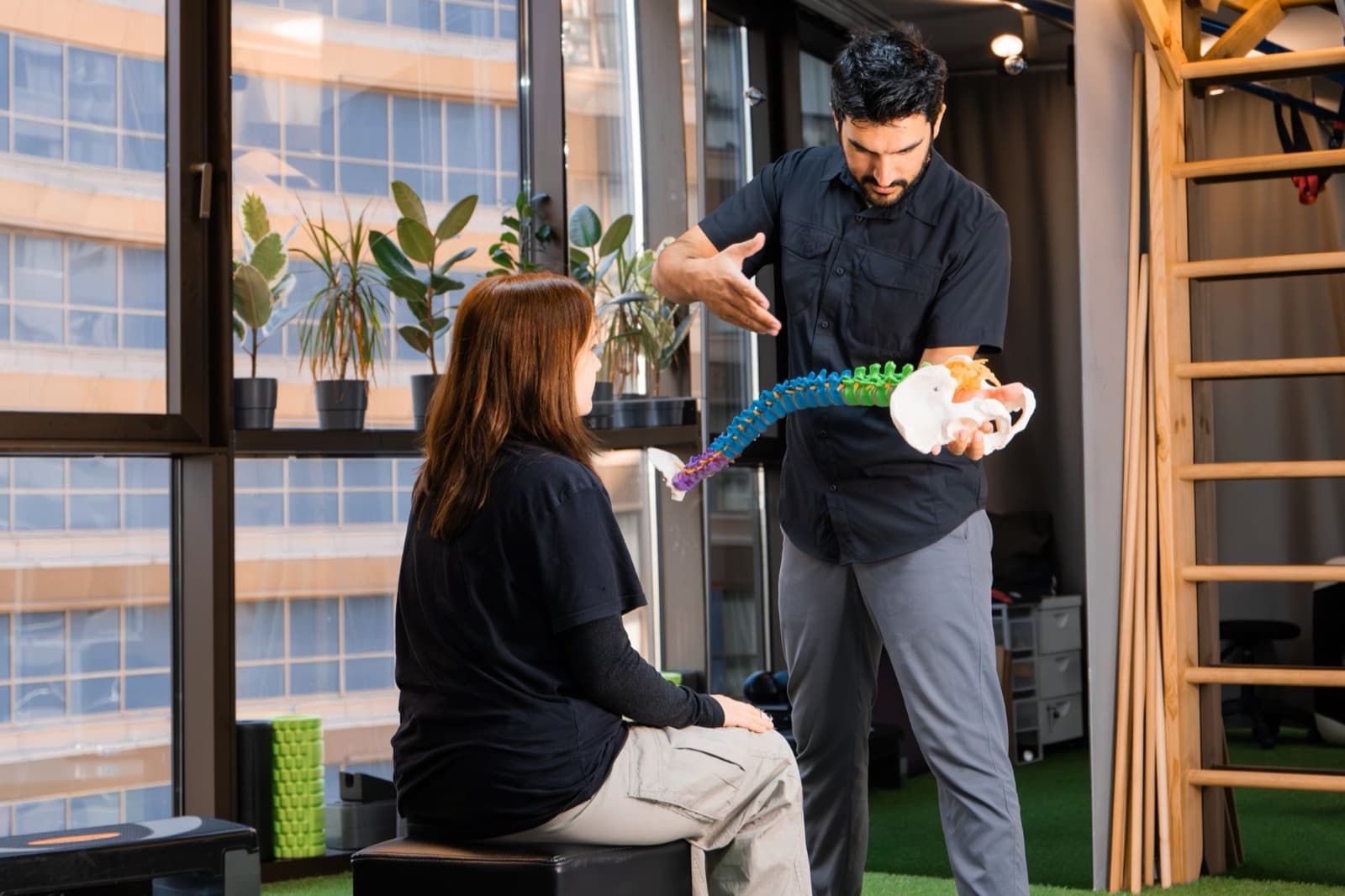 A clinician holds a color-coded anatomical spine model and explains it to a seated woman in a rehab studio with large windows and plants.