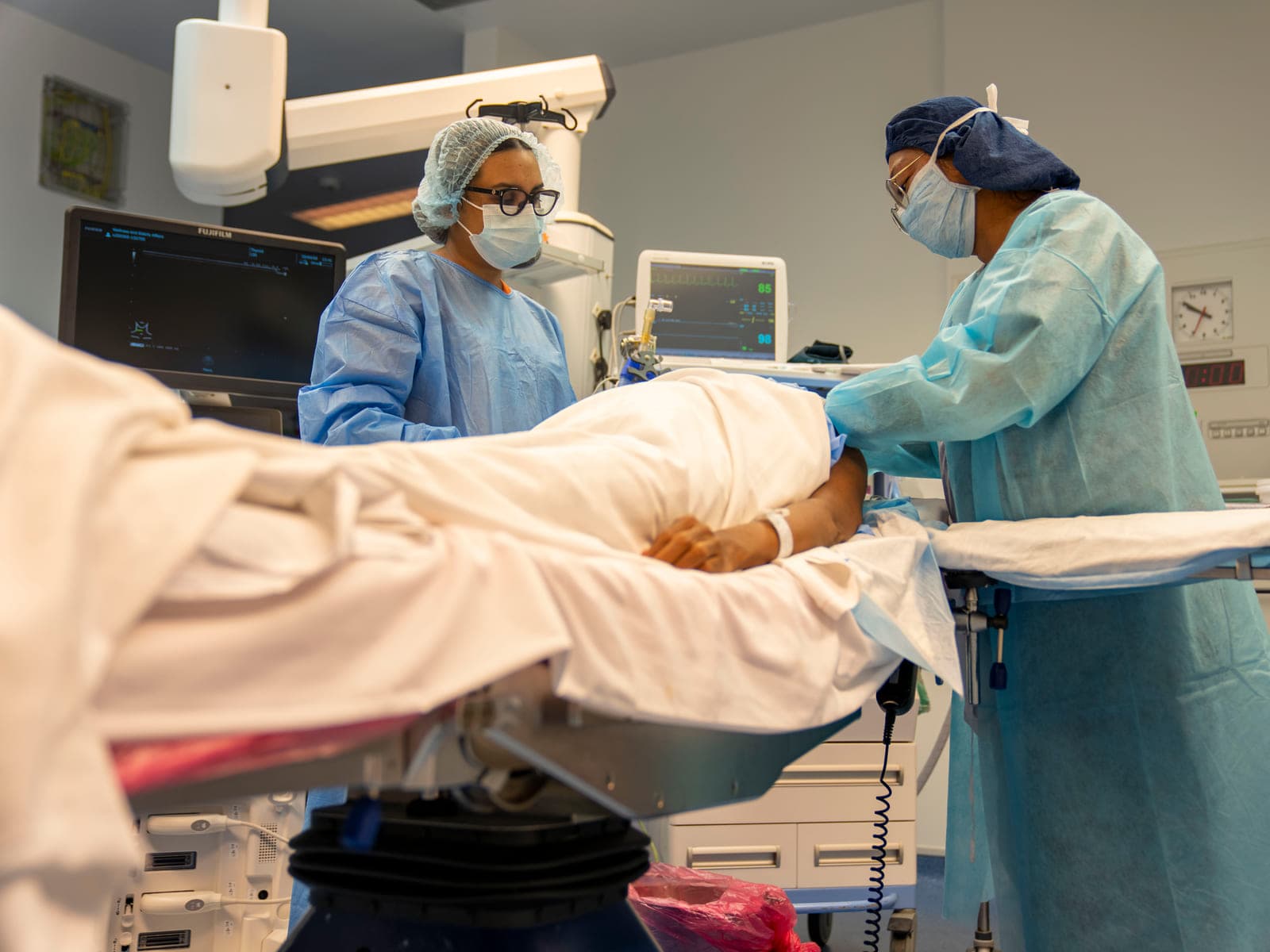 Two clinicians in surgical gowns, caps, and masks stand on either side of a patient covered by a sheet during an ultrasound-guided nerve block, an imaging monitor and a vitals monitor beside the table.