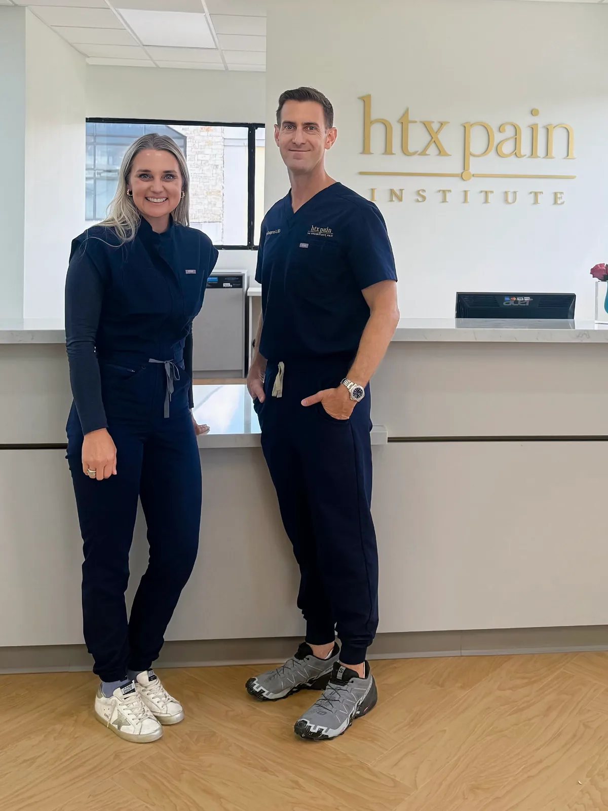 Allison Turner, DNP, and Edward Baumgartner Jr., MD, in navy scrubs, standing side by side at the clinic reception desk beneath the gold htx pain institute wall sign.