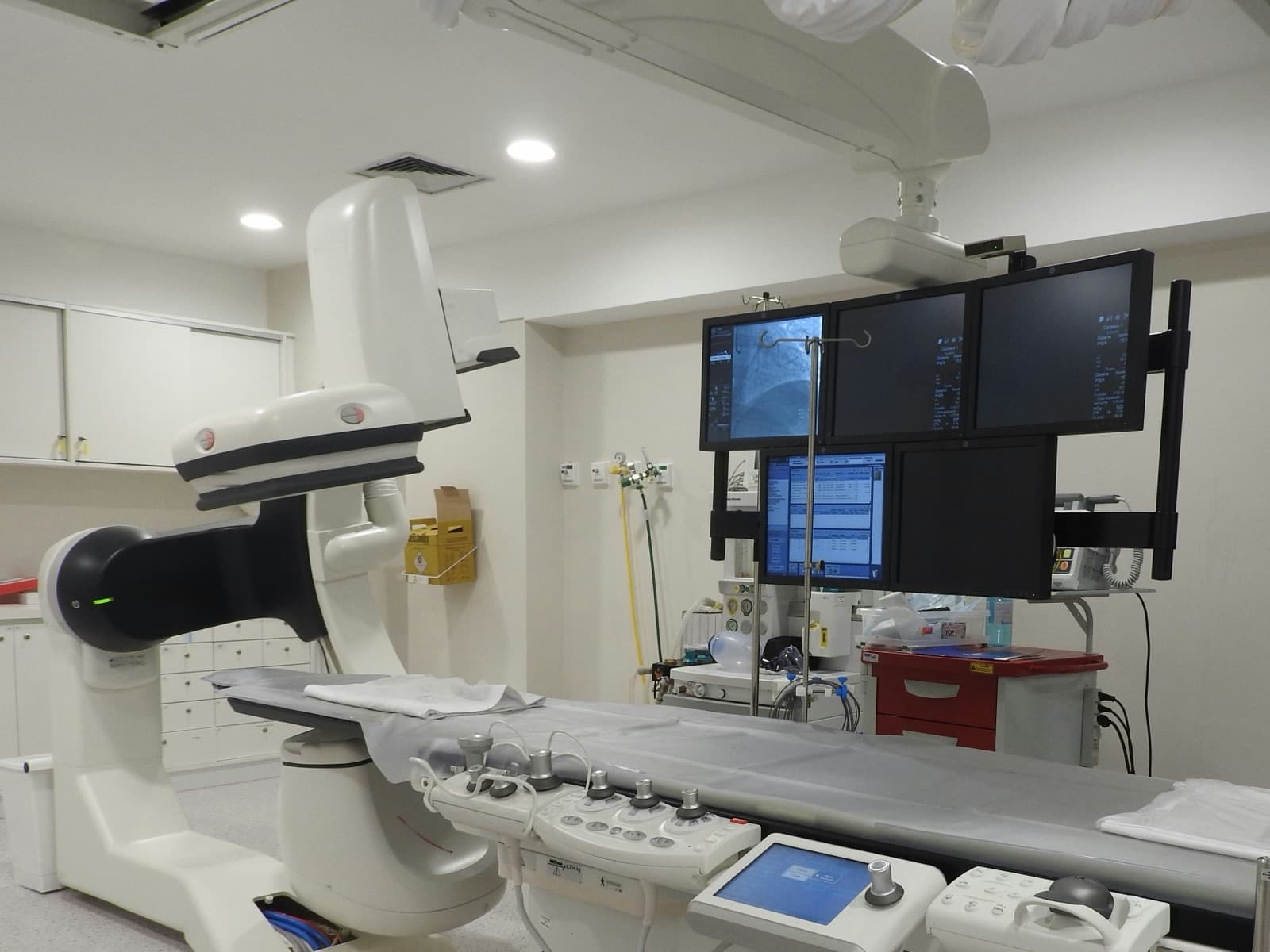 A fluoroscopy C-arm and patient table in a bright, empty interventional procedure suite with a bank of imaging monitors.
