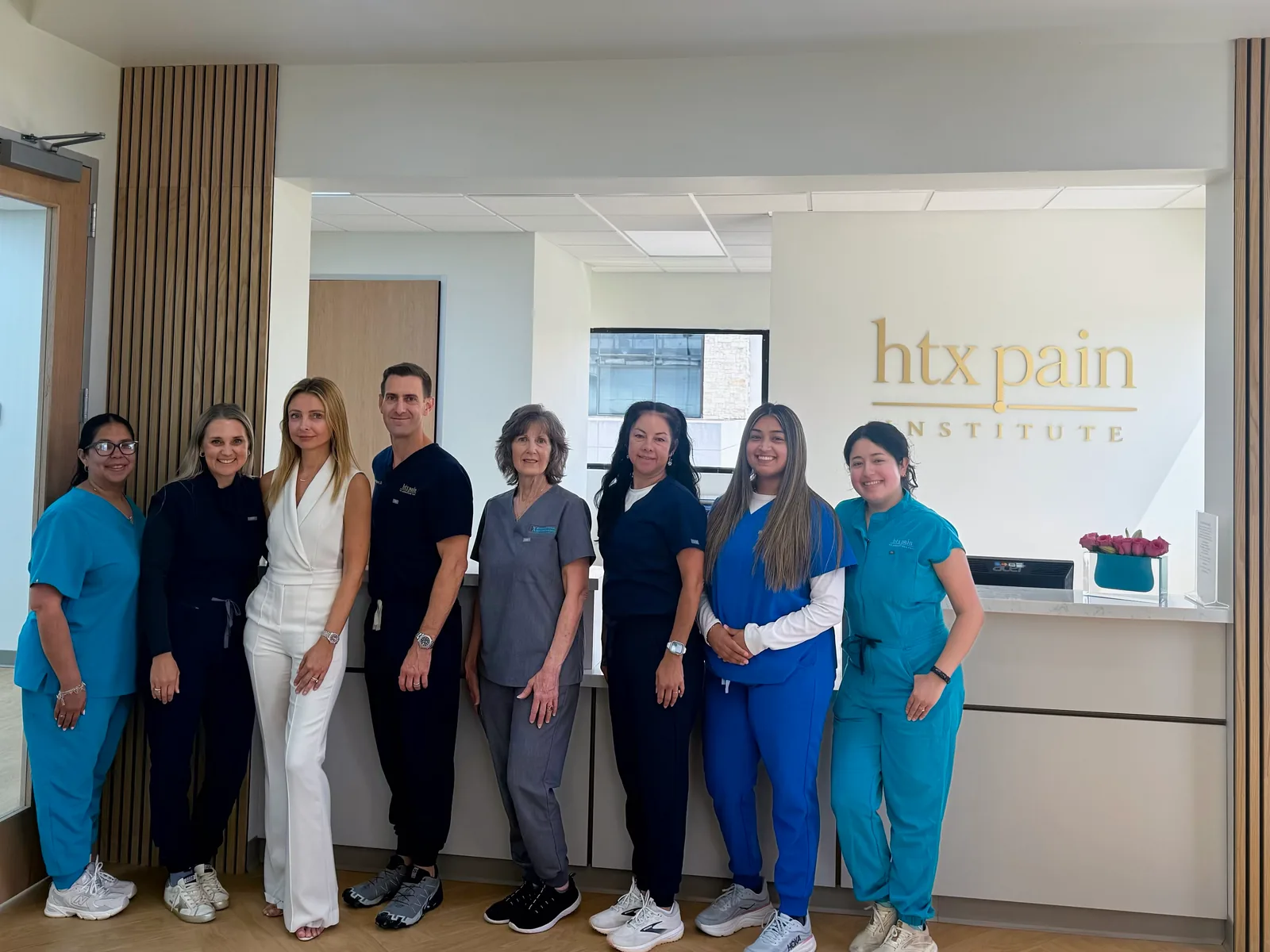 Eight team members, most in scrubs, stand shoulder to shoulder beneath the gold htx pain institute wall sign; Edward Baumgartner Jr., MD, in navy scrubs, is fourth from left.