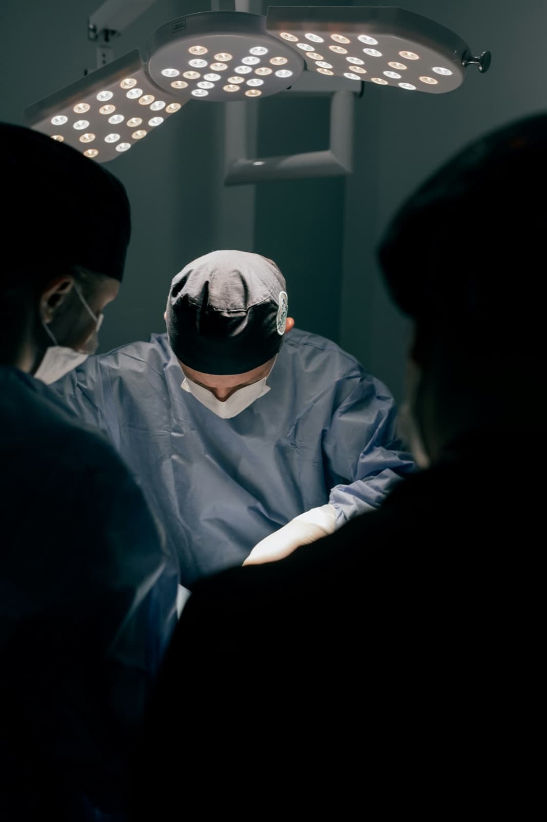 A physician in a blue surgical gown, cap, and mask looks down while working beneath bright overhead procedure lights, flanked by two colleagues seen from behind.