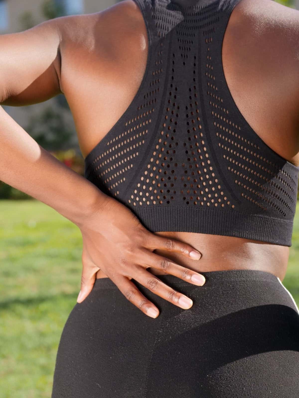 Woman in black athletic wear seen from behind outdoors, pressing one hand to her lower back.